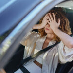 frustrated woman in car covering face with hands, wearing beige jacket and white top, sitting in driver seat with seatbelt fastened during daytime.